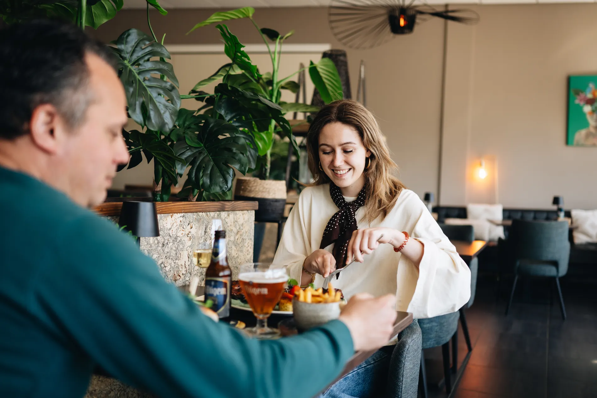 Een jonge vrouw met een sjaaltje die vrolijk dineert aan een tafel in het restaurant, met op de voorgrond een man in een groene trui en op de achtergrond grote botanische planten.